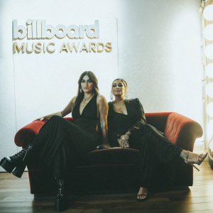 Yasmine Yousaf and Jahan Yousaf from Krewella, sitting on a red velvet couch. The backdrop features the Billboard Music Awards logo. Both sisters wear elegant black dresses. Bright stage lights illuminate the scene.