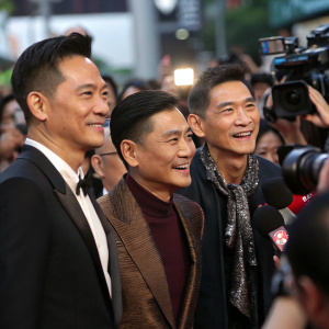 Nick Cheung, Chris Lee, Chow Yun-fat, and Andy Lau stand together at a bustling media event. Flashing cameras and microphones surround them.