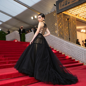 Sofia Carson at the annual Met Gala. She is wearing a long, black, intricate gown with layers of tulle and sheer fabric. The background features a modern architectural setup with gold decorations and a vibrant red curtain.