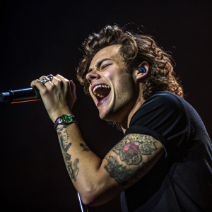 Harry Styles, the male singer, center stage with dramatic lighting. Curly hair, tattoos on arm, dark clothing, intense performance. Dark background, close-up shot.