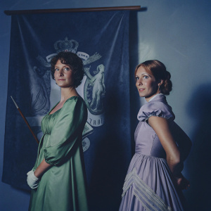 Vinda Rosier and Queenie Goldstein at the Wizarding World gala. They wear elaborate costumes, one in emerald green, the other in royal purple. Decorative backdrop features magical runes and symbols.