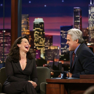 Fran Drescher, laughing heartily, alongside Jay Leno. They are in the studio audience. Backdrop features a skyline of New York City