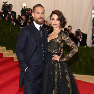 Tom Hardy and Charlotte Riley at the Met Gala. Tom Hardy in a dark suit, Charlotte Riley in a black gown with intricate lace detailing and gold accents. They stand confidently on a red carpet with paparazzi surrounding them.