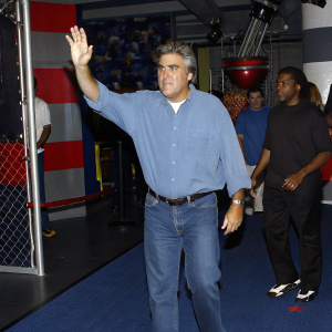 Jay Leno at the opening of a new museum exhibit. Casual attire, light blue shirt and jeans, waving to the crowd. Vibrant exhibition hall with exhibits in the background.