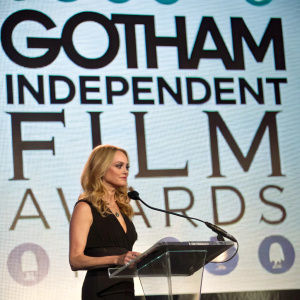 Heather Graham, in a black dress, speaking at a podium. Behind her, a large screen displaying the Gotham Independent Film Awards logo.