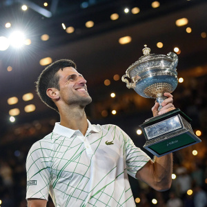 Novak Djokovic, Serbian tennis star, grinning widely. Wearing Nike shirt with crocodile emblem. Holding trophy in bright arena. Crowd blurred