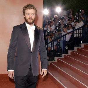 Justin Timberlake, sporting a beard and curly hair, in a tuxedo. Posing for a photo at a Hollywood event. Crowded backdrop with paparazzi and logos