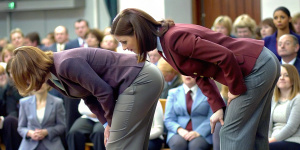 two women in their thirties with chubby bottoms and really tight bottom hugging  grey flared trousers and burgundy blazers are bending  over to be formally punished in a school assembly