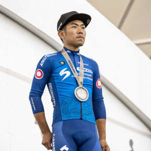 Caleb Ewan, Asian male athlete, standing tall with a stern expression. Wearing a blue cycling outfit with sponsorship logos. He holds a silver medal and wears a black cap. In front of a white background at an awards ceremony.