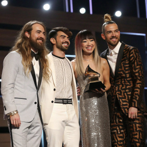 DNCE band members Jack Lawless, Joe Jonas, JinJoo Lee, and Cole Whittle posing on stage at the Grammy Awards. Jack with long hair and bow, Joe in a classic white suit, JinJoo in a sequined gown, Cole with a bold patterned outfit.
