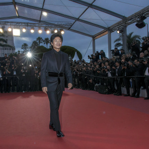Lee Byung-hun, wearing a tailored black suit and black belt, graces the Cannes Film Festival red carpet. A bustling crowd of photographers and fans in the background.