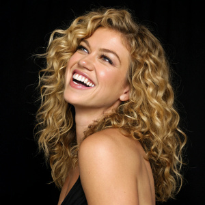 Adrianne Palicki, actress, smiling brightly. Black backdrop. Curly blonde hair