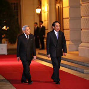 Emperor Akihito and President Tran Dai Quang in formal attire, walking down a red carpet at an international summit. Emperor with auburn hair, Caucasian features, President with East Asian appearance. Both in dark suits. Warm, golden lighting highlights the classic architecture backdrop.