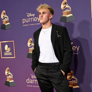Jake Paul, a young man with blonde hair, stands confidently at the Grammy Awards. He is wearing a black blazer and white t-shirt. The backdrop features Disney logos and decorative items. The purple background contrasts beautifully with his outfit.