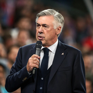 Carlo Ancelotti, wearing a dark suit and tie, addressing the press conference. Intense facial expression. A blurred audience in the background.