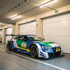 The Audi RS5 DTM car, with blue and white paint job, green and yellow accents, sitting in a garage. Neatly parked with sponsor logos visible. Neutral colored walls