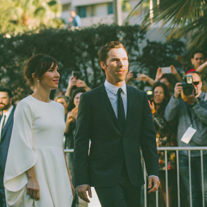 Benedict Cumberbatch and Sophie Hunter on the Cannes Film Festival red carpet. Cumberbatch in a tailored black suit and tie, Hunter in a stunning white dress. Crowded backdrop with flashing cameras and enthusiastic fans.