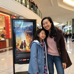 Christy Chung and her daughter Cayla Janie Yen smiling in a shopping mall, standing next to movie poster for upcoming blockbuster.