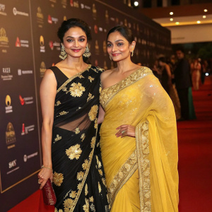 Pooja Kumar and Andrea Jeremiah at a luxury gala. Both in stunning traditional Indian attire. Pooja in black saree with gold floral patterns, Andrea in yellow saree with intricate detailing. Red carpet ambiance, branded backdrop visible.
