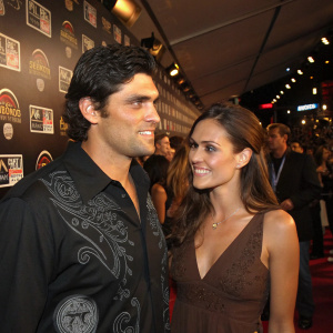 Mark Philippoussis and Amanda Salinas mingle at a crowded gala. Mark, with dark hair and a black shirt featuring a graphic design, stands alongside Amanda, who is wearing a brown dress. The backdrop is a chaotic wall filled with various logos and signage.