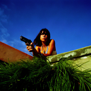 Mathilda, from Léon: The Professional, in a tense pose. Dark hair, serious expression, wearing a necklace. Shot against a stark black backdrop.