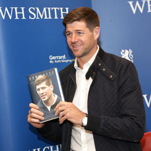 Steven Gerrard, former soccer player, holding a copy of his autobiography 'Gerrard My Autobiography' at a WH Smith promotional event. Wearing a dark jacket and crisp white shirt. Against a solid blue background with WH Smith logos and text.