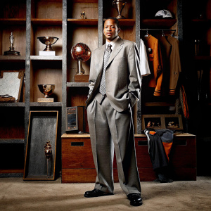 Allen Iverson at an NBA hall of fame induction. Dressed in a tailored suit, Iverson poses confidently in a dimly lit room filled with sports memorabilia.