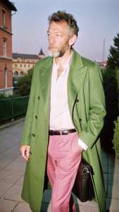In the photo, Italian actor Vincent Cassel strolls through Milan Fashion Week in a dark green oversize coat, pale pink trousers, a white shirt and black loafers, a black bag hangs in his hands, a slight smile on his face, black hair with gray and careless stubble, he is depicted completely tall, his gaze is not directed at the camera, he passes by the camera, the object is on the shoulder side, the city is in the background