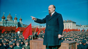 Generate a photorealistic image of Vladimir Lenin delivering a speech at a rally in the early 20th century. Lenin is standing on a wooden platform, passionately addressing a large crowd. He is wearing a dark suit, a peaked cap, and is gesturing emphatically. The background includes a sea of people waving red flags and banners, with historical buildings of early 20th-century Russia in the backdrop. The scene is lit by natural daylight, capturing the fervor and energy of the moment.
