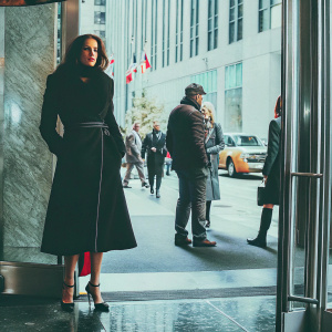 Eva Green standing confidently outside SiriusXM Studios. She wears a sleek black coat and high heels. The backdrop is the bright, bustling city street.