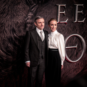 Martin Freeman and Amanda Abbington at a premiere. He wears a black suit, she in a white blouse and black skirt. Dark and dramatic backdrop with large EE logo