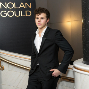 Nolan Gould, a young actor with curly dark hair, is photographed in a formal setting. He wears a black suit and white shirt. The background is a sleek, modern design with a logo.