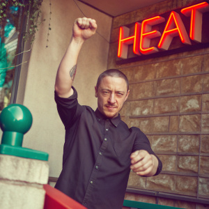 James McAvoy, white male with short hair, striking a pose. Wears a black button-down shirt with a visible tattoo on his forearm. Standing in front of a vibrant 'Heat' billboard and a modern brick wall backdrop.