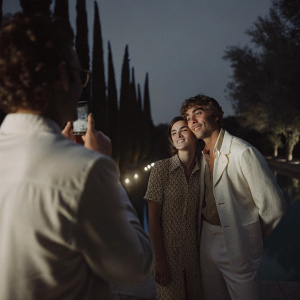 Ross Lynch and Kiernan Shipka posing for a candid photo. Roberto Aguirre-Sacasa stands beside them. Soft evening lighting, surrounded by tall trees and flickering candles.