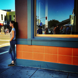 Emmy Rossum, in a casual outfit, standing alone at a cafe. Background is bustling street life.