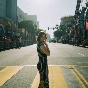 Lauren Cohan in a black and white striped dress. Short brown hair. The backdrop is a brightly lit, sunny street scene. She's posing for a fan at Comic-Con.