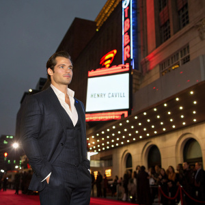 Henry Cavill at premiere event, wearing a dark suit and crisp white shirt. The background showcases a large billboard with the movie title in neon lights.