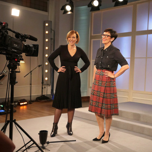 Mel Giedroyc and Sue Perkins on the set of a talk show. Mel in a elegant black dress, Sue in a stylish plaid skirt and blouse.