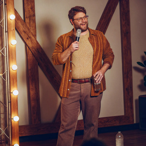 Seth Rogen, sporting a casual outfit and beard, holding a microphone and water bottle. Brightly lit stage backdrop.