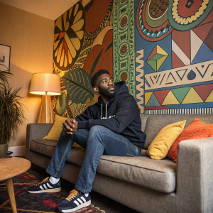 Tinie Tempah, in a cozy living room setting. He's casually dressed in a dark hoodie and jeans. A vibrant mural wall with geometric patterns and bright colors serves as the backdrop.
