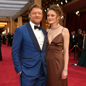 Sean Bean and Ashley Moore at a glamorous gala event. Sean in a blue tuxedo, Ashley in a brown evening gown. Red carpet backdrop.