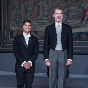 Carlos Alcaraz and King Felipe VI at a grand gala. Carlos in dark blazer, King in traditional suit. Both smiling. Behind them, a detailed tapestry with vibrant figures and colors.