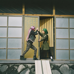 Jolyne and Anna Sui in a vibrant comic panel from 'Jojo's Bizarre Adventure - Stone Ocean'. Jolyne's long pink hair contrasts with Anna Sui's green hair against a bright yellow backdrop with a graphic pattern. The illustration captures a dynamic, dramatic scene.