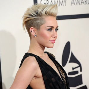 Close-up shot of Miley Cyrus at award show. She wears a sleek black gown with intricate details, paired with an edgy short haircut. The backdrop is plain white with subtle lighting creating a stark contrast.