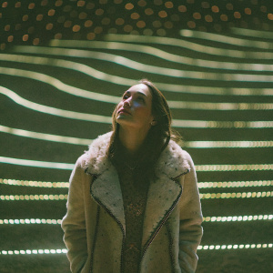 Claire Forlani, an actress, with a serene expression. Wearing a beige fur coat with intricate detailing. The background is an abstract pattern of blurred lights.