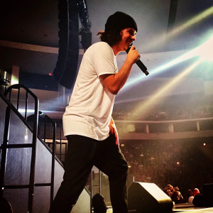 Lukas Graham Forchhammer on stage at the 2016 Jingle Ball. Wearing a casual white t-shirt and black beanie, he performs with passion under dramatic lighting. Stage equipment and crowd visible.