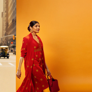 Freida Pinto, Indian actress, in a vibrant red dress with intricate gold embroidery. Walking on a busy city street with passing pedestrians and vehicles.