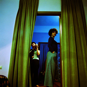 Vintage photo from the 1920s. Woman with short curly hair, standing in a room with a curtain. Wearing a dark top and long loose-fitting skirt. Another person taking the photo.