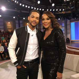 Jonathan 'Heat' Martinez and Vivica A. Fox on a talk show set. Jonathan is in a white shirt, black jacket, and black pants. Vivica is in a black sequined dress. Bright lights and audience visible