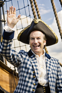 Bob Odenkirk in role of pirate salesman Stan from "Monkey island",with a big bright smile, waving his arms, in a blue checkered jacket, pirate shp in background
